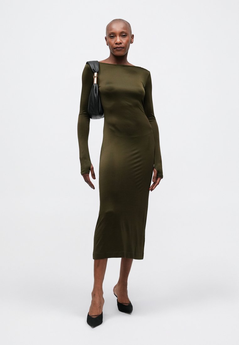 Robe longue à manches longues de couleur vert olive avec une silhouette ajustée, présentant un col bateau. Le modèle tient un sac à main noir et porte des talons aiguilles noirs.