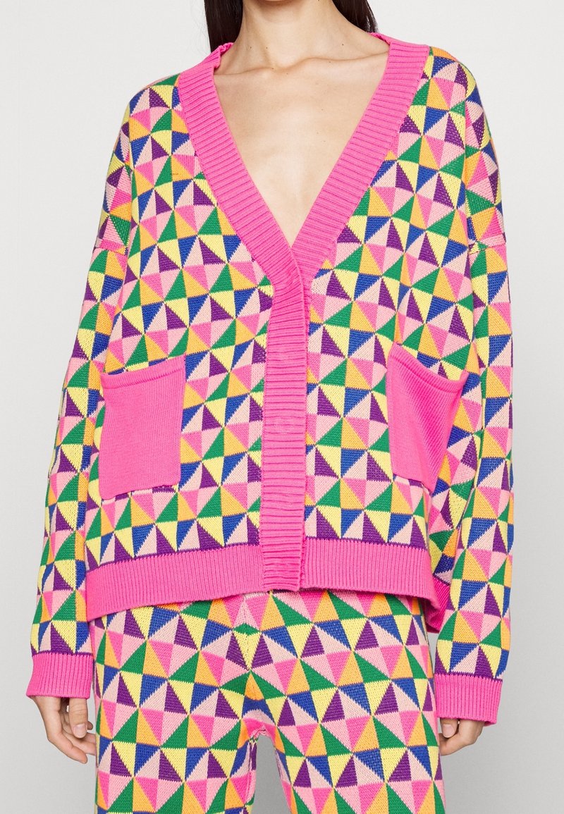 Femme portant un cardigan en maille coloré et un pantalon avec un bord rose et un motif géométrique triangulaire multicolore en jaune, vert, bleu, violet et rose.