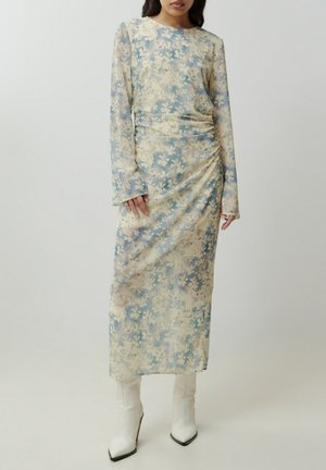 Ein blumendrucktes Kleid aus weichem, transparentem Stoff in Blau- und Cremetönen, mit figurbetontem Schnitt und gerafften Details, kombiniert mit weißen Absatzstiefeln.