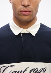 Cămașă polo bleumarin cu guler alb și clapetă cu nasturi. Prezintă o bandă albă supradimensionată cu text bleumarin pe piept. Material din bumbac.