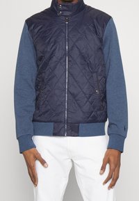 Gewatteerde navy voorkant met blauwe mouwen, ritsluiting, hoge kraag, geribbelde manchetten en zoom. Linker ritszak; subtiel logo op de manchetten.