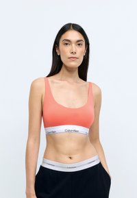 Korall színű sportmelltartó széles pántokkal és mély kivágással, fehér szalaggal, amelyen a "Calvin Klein" logó látható. Sima textúra; minimalista design.
