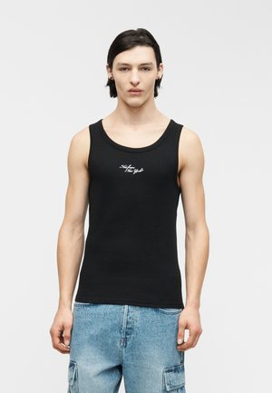 Giovane uomo con capelli lisci e scuri indossa una canottiera nera con scritta bianca e pantaloncini cargo in denim azzurro, in piedi davanti a uno sfondo neutro.