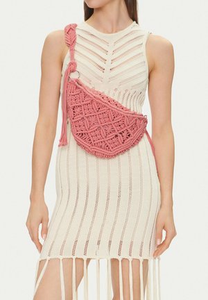 Frau trägt ein ärmelloses, cremefarbenes Strickkleid mit vertikalen Mustern und eine rosa Makramee-Umhängetasche mit Fransen.