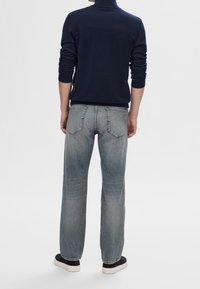 Man som bär urblekta raka jeans, marinblå tröja och svarta sneakers, står vänd bort mot en vit bakgrund.