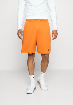 Hombre vestido con pantalones cortos deportivos de color naranja brillante con el logo negro de Nike, camiseta blanca de manga larga, calcetines blancos de Nike y zapatillas blancas sobre un fondo neutro.