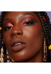 Närbild av ett ansikte med flätat hår, djärv röd och mörk ögonsminkning, en matt brun läppfärg och stora, utsmyckade guldfärgade örhängen.