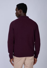Pull maroon avec un col polo, tissu texturé, manches ajustées et un ourlet droit. Porté avec un pantalon beige, vu de dos.