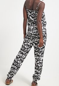 Svart och vit jumpsuit med palmbladsdesign, spagettistilar, avslappnad passform och elastisk midja. Tyget verkar lätt och slätt.