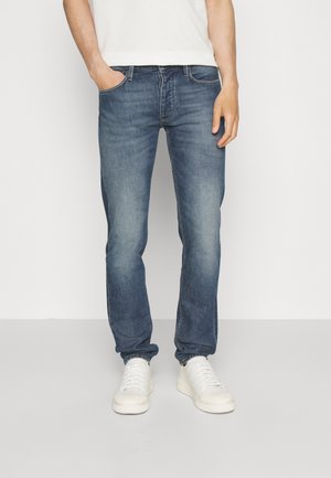 Mann trägt blaue, ausgebleichte Denim-Jeans und weiße Turnschuhe, steht mit einer Hand in der Tasche vor einem schlichten Hintergrund.