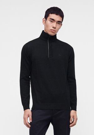 Jeune homme portant un pull noir à manches longues avec un col zippé montant et un pantalon foncé, debout devant un fond clair uni.