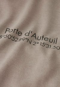 Smėlio spalvos audinys su užrašu „Porte d'Auteuil“ ir geografinių koordinačių spausdinimu. Lygus paviršius, aiškus juodas šriftas, artimas vaizdas.