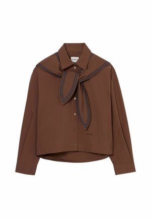 Giovane donna che indossa una camicia marrone con sciarpa, pantaloni beige a gamba larga e scarpe marrone scuro, in piedi davanti a uno sfondo chiaro e semplice.