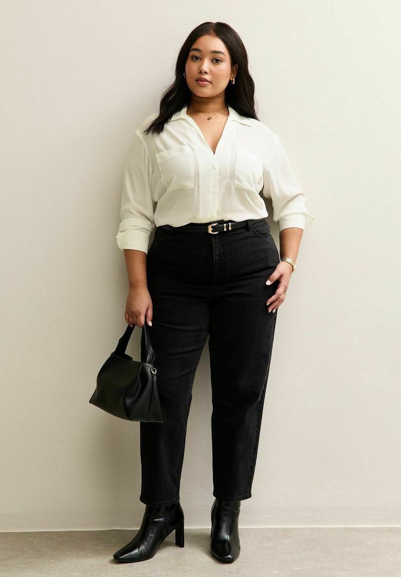 Kvinna står mot en vägg iklädd vit blus, svarta jeans med hög midja, svarta stövlar och håller en svart handväska i vänster hand.