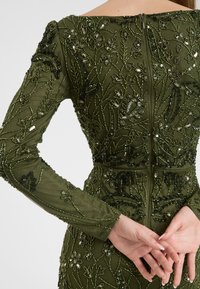 Abito verde a maniche lunghe con delicata ricamo di perline e fiori. Presenta una silhouette aderente e una cerniera nascosta sul retro per la chiusura.