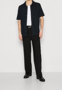 Schwarz kurzärmeliges Hemd mit Kragen und Knöpfen, getragen über einem weißen Unterhemd, kombiniert mit schwarzen Hosen und schwarzen Schuhen. Einfaches Design, strukturierter Stoff.