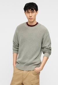 Νεαρός άντρας που κοιτάζει μπροστά, φορώντας ένα ανοιχτό γκρι πλεκτό πουλόβερ πάνω από ένα μπορντό πουκάμισο, με τα χέρια στις τσέπες των ταμπά cargo παντελονιών, με φόντο λευκό.