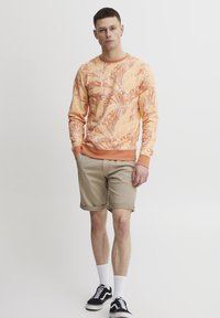 Sudadera naranja con estampado de hojas, puños y cuello acanalados. Pantalones cortos beiges con dobladillos enrollados. Zapatillas negras con suelas blancas, calcetines blancos.