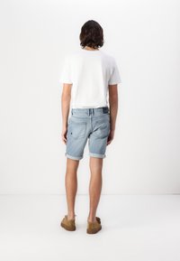 Muž stojící zády, oblečený v bílém tričku, světlých modrých džínových kraťasech s ohrnutými nohavicemi a hnědých volnočasových botách na jednobarevném bílém pozadí.