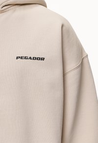 Beige bomullströja med avslappnad passform, ribbade muddar och en svart "PEGADOR"-logotyp på bröstet. Mjuk textur och enkel design.