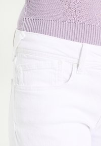 Bílé denimové džíny s úzkým střihem, předními a zadními kapsami a jemnými šicími detaily. Hladká textura, rovný střih nohavic.