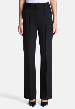 Femme portant un pantalon noir taille haute à jambes larges avec une ceinture noire assortie et des chaussures noires à bout pointu, debout sur un fond uni.