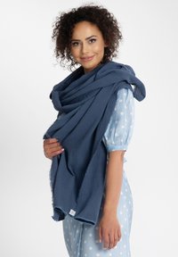 Teksturowany, ciemnoniebieski szalik wykonany z lekkiego materiału, swobodnie zarzucony na szyję, z surowymi brzegami i widoczną małą metką.