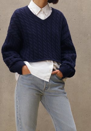 Mujer con un suéter corto de punto con trenza azul marino sobre una camisa blanca abotonada metida en unos vaqueros azul claro, con las manos en los bolsillos.