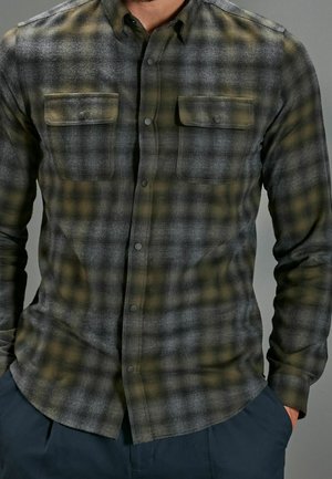 Mann trägt ein grünes und graues kariertes Hemd mit Knöpfen und zwei Brusttaschen, kombiniert mit einer dunklen Hose, Hände in den Taschen.