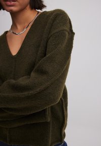 Persoon in een olijfgroene fuzzy gebreide V-hals trui en een zilveren ketting, weergegeven van schouders tot halverwege de taille.