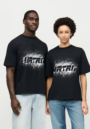 Deux mannequins posent côte à côte, vêtus de T-shirts noirs à motifs graphiques et de jeans, devant un fond clair et uni.