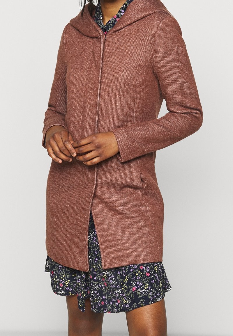 Cappotto marrone con cappuccio e finitura strutturata, maniche lunghe e tasche laterali, abbinato a un abito floreale blu con orlo arricciato.