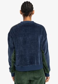 Felpa in velluto blu navy con pannelli laterali verdi. Presenta polsini e scollo a coste, e dettagli metallici lungo la cucitura della spalla.