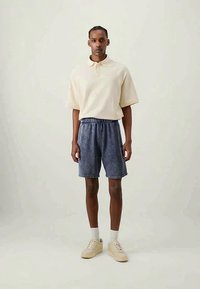 Man standing wearing a cream short-sleeve polo, blue elastic waist shorts, white socks, and beige sneakers on a plain light background.