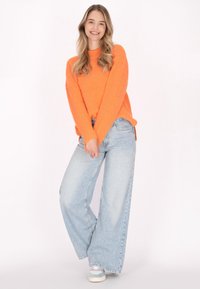 Helles orange gestricktes Sweatshirt mit langen Ärmeln, kombiniert mit hellblauen weiten Jeans und mehrfarbigen Sneakers. Der Hintergrund ist schlicht weiß.