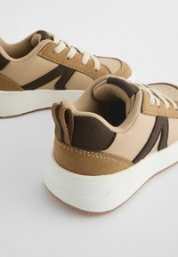 Baskets beige et marron avec une tige texturée, des accents contrastants et des semelles en caoutchouc blanc. Présentent des lacets ronds et des détails perforés.