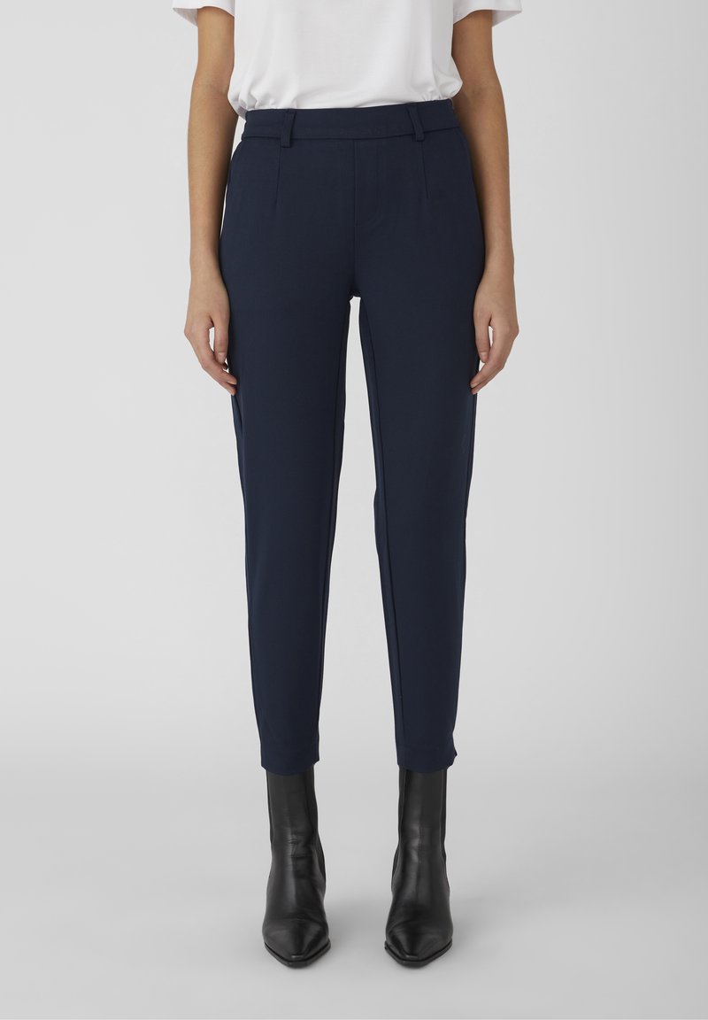 Femme portant un pantalon slim bleu marine sur mesure et des bottines noires à bouts pointus, debout devant un fond gris clair uni.
