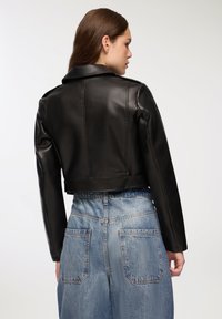 Giubbotto in pelle nera tagliato con texture liscia, colletto ampio e cuciture sulle spalle. Abbinato a pantaloni in denim alta vita di un blu chiaro.