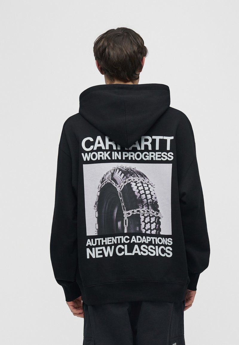 Persona che indossa una felpa nera con ampio testo bianco e immagine di pneumatico con catene sul retro, in piedi davanti a uno sfondo neutro.