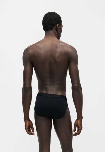 Άνδρας που στέκεται με την πλάτη, φοράει μαύρα σλιπ, με απλό ανοιχτό γκρι φόντο.