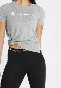 Camiseta de algodón gris con detalle lateral anudado y logo blanco de "Champion". Combinada con leggings negros que tienen una cinturilla con el mismo logo.