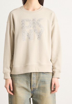 Beige Sweatshirt mit "DKNY" in silbernen Perlen buchstabiert, getragen zu hellblauen, ausgewaschenen Jeans, gezeigt vom Hals bis zur Hüfte.