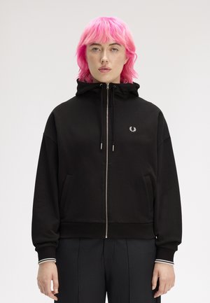 Personne aux cheveux rose vif portant un sweat à capuche noir zippé avec un petit logo de laurier blanc sur la poitrine, sur un fond uni.