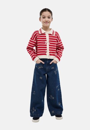 Jeune fille portant un pull boutonné rouge et blanc à rayures et un jean large bleu foncé avec des motifs brodés blancs, debout les mains dans les poches.