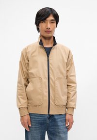 Ανακριβές μπεζ επενδυμένο μπουφάν με όρθιο γιακά, μπροστινό κλείσιμο με φερμουάρ, δύο μεγάλες τσέπες και  ribbed μανσέτες, φορεμένο πάνω από μια ριγέ μπλούζα.