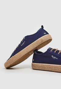 Námořnické plátěné tenisky s texturovanou, tkanou juty podrážkou a hnědými tkaničkami. Obsahují logo na boční straně a vzorovaný detail na patě.