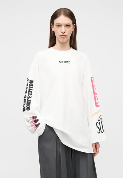 Γυναίκα που φοράει λευκό πουκάμισο μακρυμάνικο με την επιγραφή "STIEGLITZ" στο στήθος και σχέδια με κείμενο και στα δύο μανίκια, σε συνδυασμό με σκούρο γκρι παντελόνι.
