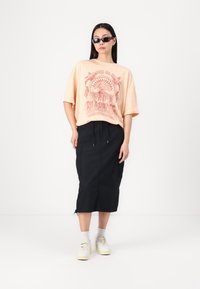 Peachfarvet oversized t-shirt med grafisk print, parret med en sort midinederdel med sidelommer og snøre. Hvide sneakers afslutter looket.