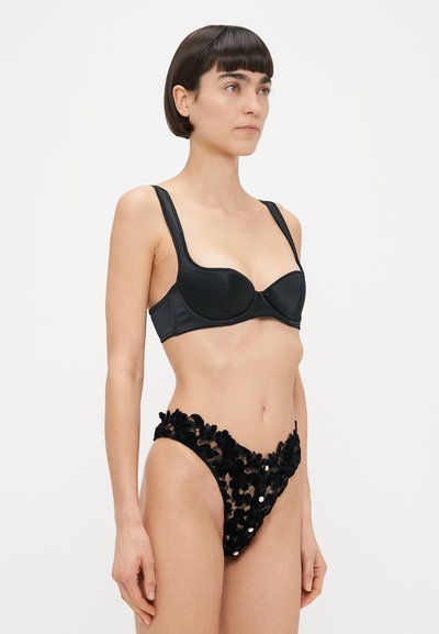 Femme aux cheveux courts et foncés portant un soutien-gorge en satin noir et un shorty assorti en dentelle florale transparente, debout devant un fond blanc uni.