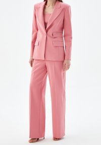 Kobieta w różowym, dopasowanym garniturze z szerokimi spodniami, pasującą marynarką i beżowymi, odkrytymi czółenkami na tle białego tła.
